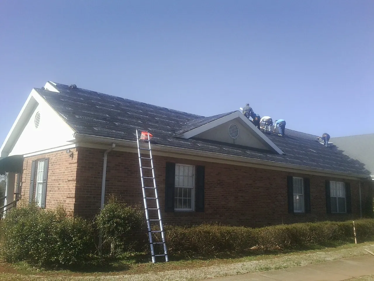 Crew tearing shingles off a brick ranch roof with a ladder set — Emergency Roof Repair work by our crew in Lime Ridge
