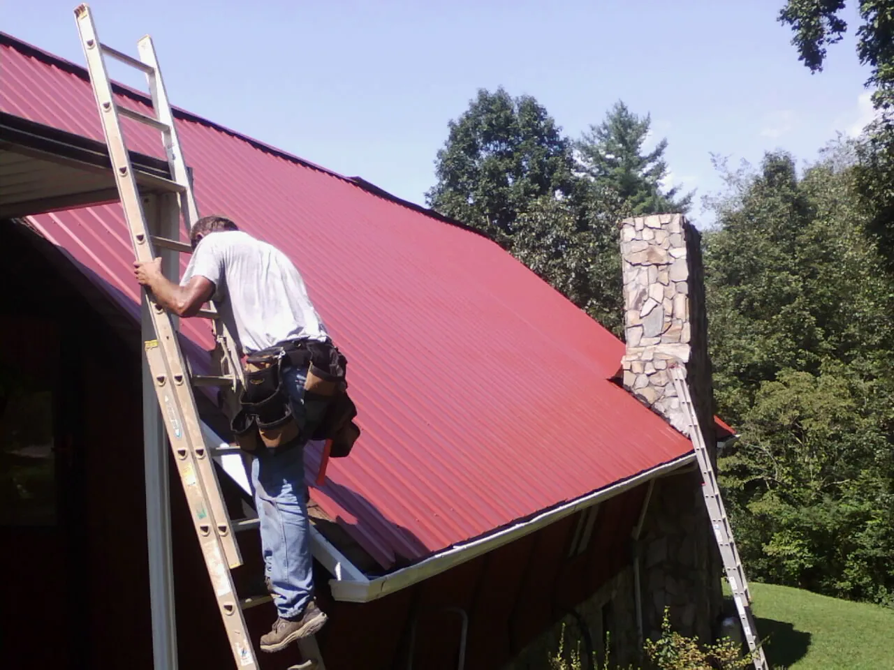 Roofer on a ladder beside a red metal roof and stone chimney — Soffit & Fascia Repair by Roofing Brothers in Lime Ridge